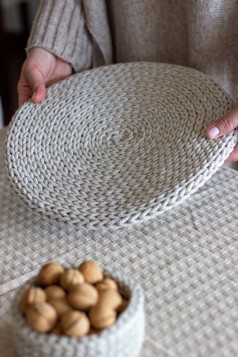 May include: A close-up of a beige crocheted round placemat being held by a hand. The placemat is made of thick rope and has a circular pattern. The placemat is sitting on a beige tablecloth.