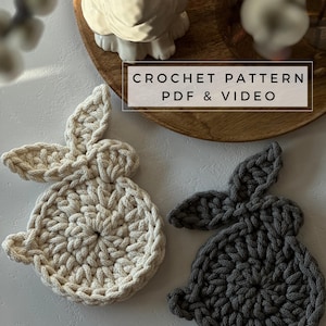 May include: Two crocheted bunny coasters, one in beige and one in gray. The coasters are on a white surface. The text "CROCHET PATTERN PDF & VIDEO" is on a wooden surface in the background.