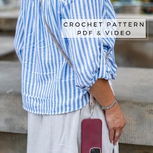 Puede incluir: Una persona lleva una camisa a rayas azules y blancas con una correa de teléfono de crochet unida a una funda de teléfono burdeos. El texto "CROCHET PATTERN PDF & VIDEO" se muestra en la imagen.