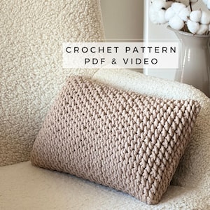 May include: A light brown crochet cushion with a textured surface. The cushion is placed on a white, textured surface. Text on the image reads "CROCHET PATTERN PDF & VIDEO".