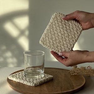 K&ouml;nnte beinhalten: Ein quadratischer, cremefarbener H&auml;keluntersetzer liegt auf einem Holztablett, mit einem klaren Glas und einem weiteren Untersetzer in der N&auml;he. Eine Hand h&auml;lt einen passenden Untersetzer dar&uuml;ber. Die Untersetzer haben ein strukturiertes Aussehen.