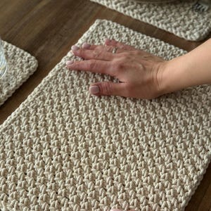 K&ouml;nnte beinhalten: Cremefarbene, handgeh&auml;kelte Tischsets und ein Untersetzer. Die Tischsets haben ein strukturiertes, gewebtes Aussehen. Das Bild zeigt die Tischsets auf einem Holztisch, mit einer Hand, die auf einem davon ruht. Der Untersetzer ist links unten sichtbar.