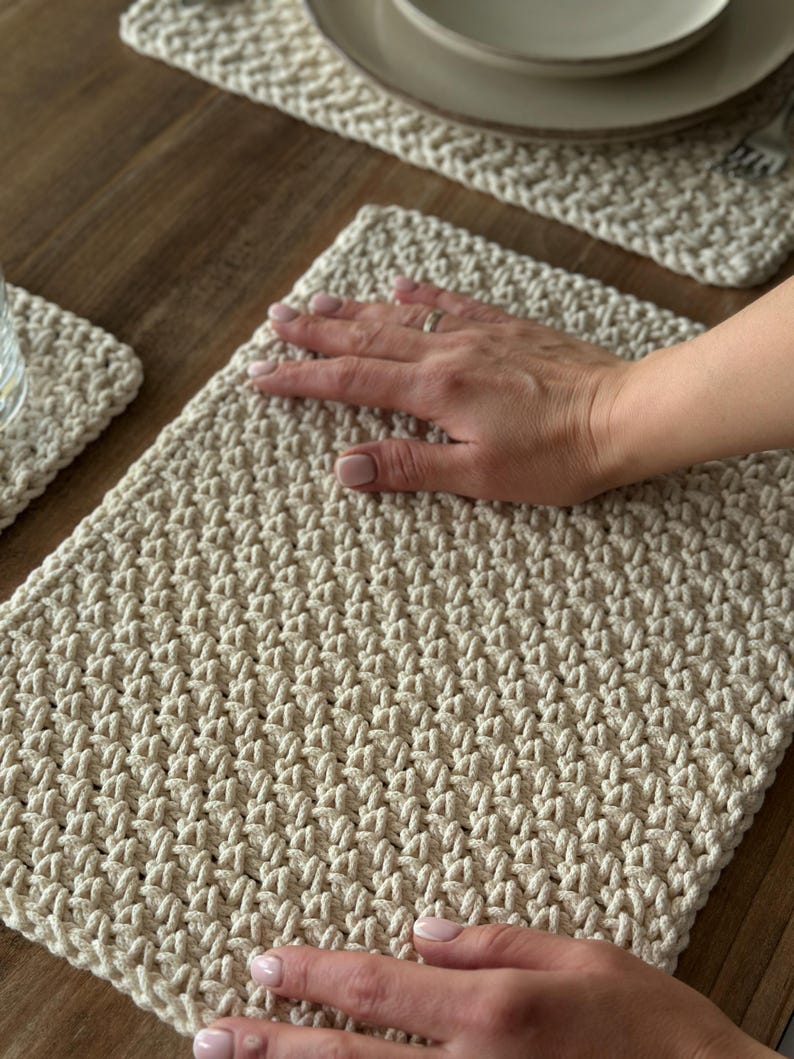 K&ouml;nnte beinhalten: Cremefarbene, handgeh&auml;kelte Tischsets und ein Untersetzer. Die Tischsets haben ein strukturiertes, gewebtes Aussehen. Das Bild zeigt die Tischsets auf einem Holztisch, mit einer Hand, die auf einem davon ruht. Der Untersetzer ist links unten sichtbar.