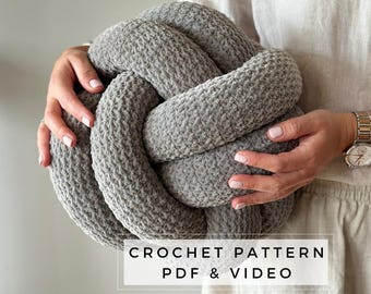 Puede incluir: Un cojín de nudo tejido a crochet gris con una superposición de texto que dice "CROCHET PATTERN PDF & VIDEO".