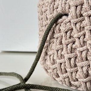 May include: A close-up of a beige crocheted bag with a green cord threaded through the top. A gold crochet hook is lying on a white surface next to the bag.