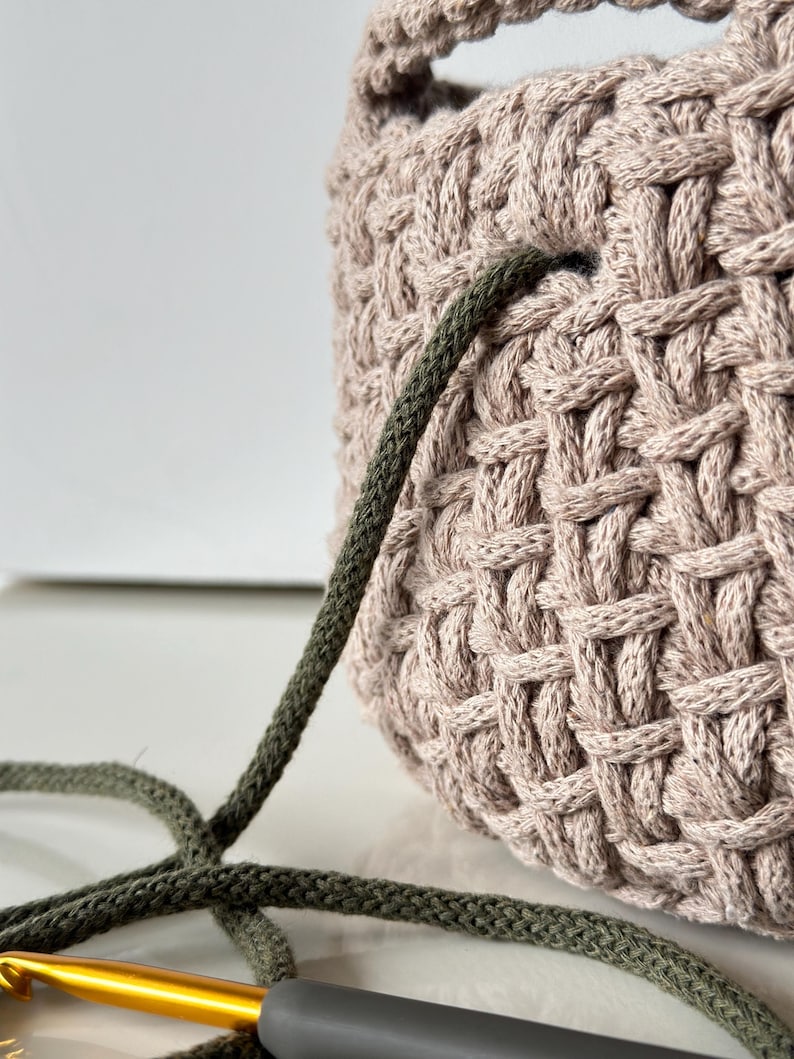 May include: A close-up of a beige crocheted bag with a green cord threaded through the top. A gold crochet hook is lying on a white surface next to the bag.