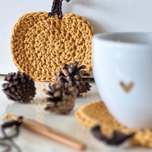 May include: A set of four crocheted pumpkin-shaped coasters in a mustard yellow color with a brown crocheted stem. The coasters are displayed with a white mug and pine cones.