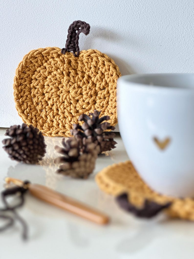 May include: A set of four crocheted pumpkin-shaped coasters in a mustard yellow color with a brown crocheted stem. The coasters are displayed with a white mug and pine cones.