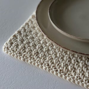 May include: A cream-colored, hand-crocheted placemat with a textured pattern. Two beige ceramic plates with a brown rim are placed on top. The placemat and plates are on a white surface.