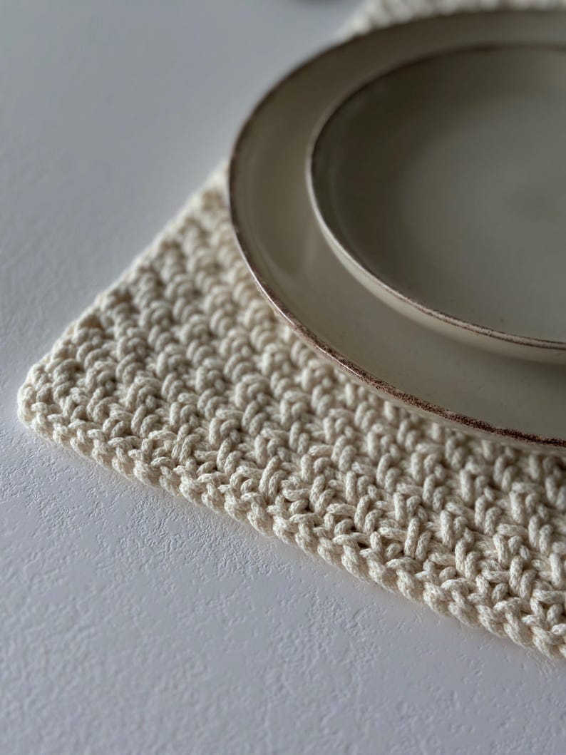 May include: A cream-colored, hand-crocheted placemat with a textured pattern. Two beige ceramic plates with a brown rim are placed on top. The placemat and plates are on a white surface.