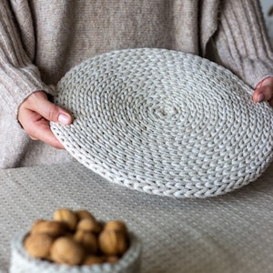 May include: A close-up of a crocheted round coaster in a light beige color. The coaster is made of thick yarn and has a circular pattern. The coaster is being held by a person's hands.