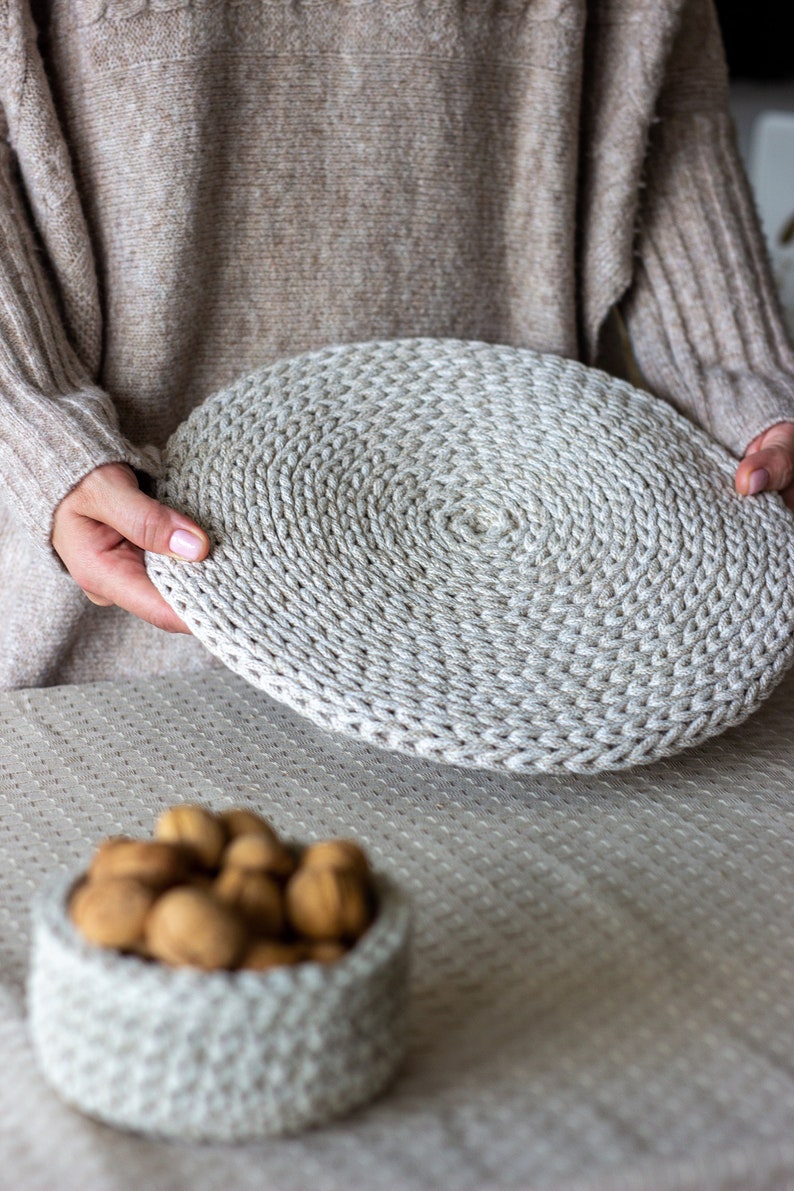 May include: A close-up of a crocheted round coaster in a light beige color. The coaster is made of thick yarn and has a circular pattern. The coaster is being held by a person's hands.
