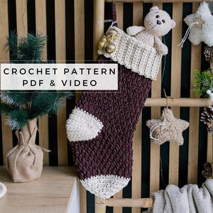 Può includere: Un calzino di Natale all'uncinetto di colore marrone e bianco. Il calzino è appeso a una scala di legno con un orsacchiotto che spunta dalla parte superiore. Il testo "CROCHET PATTERN PDF & VIDEO" è visualizzato nella parte inferiore dell'immagine.