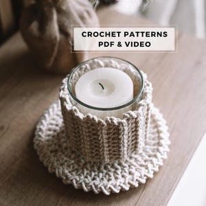 Pode incluir: Um castiçal de crochê creme com uma inserção de vidro segurando uma vela branca. O castiçal fica em uma base de crochê combinando. A imagem inclui o texto "CROCHET PATTERNS PDF & VIDEO".