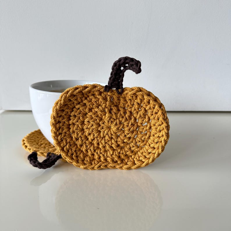 May include: Two crocheted pumpkin-shaped coasters in a mustard yellow color with brown stems. The coasters are on a white surface.
