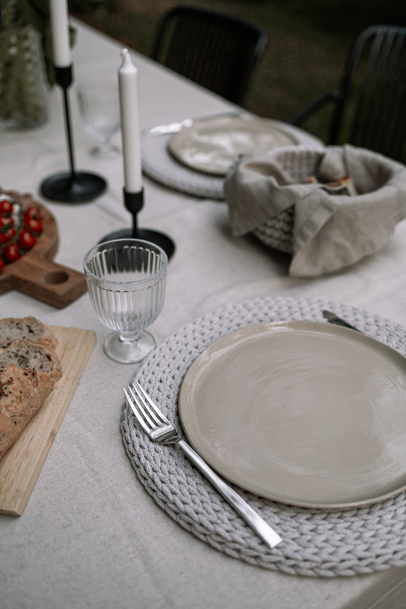 May include: A close-up of a table setting with a white tablecloth, a gray crocheted placemat, a beige plate, a silver fork, and a clear glass.