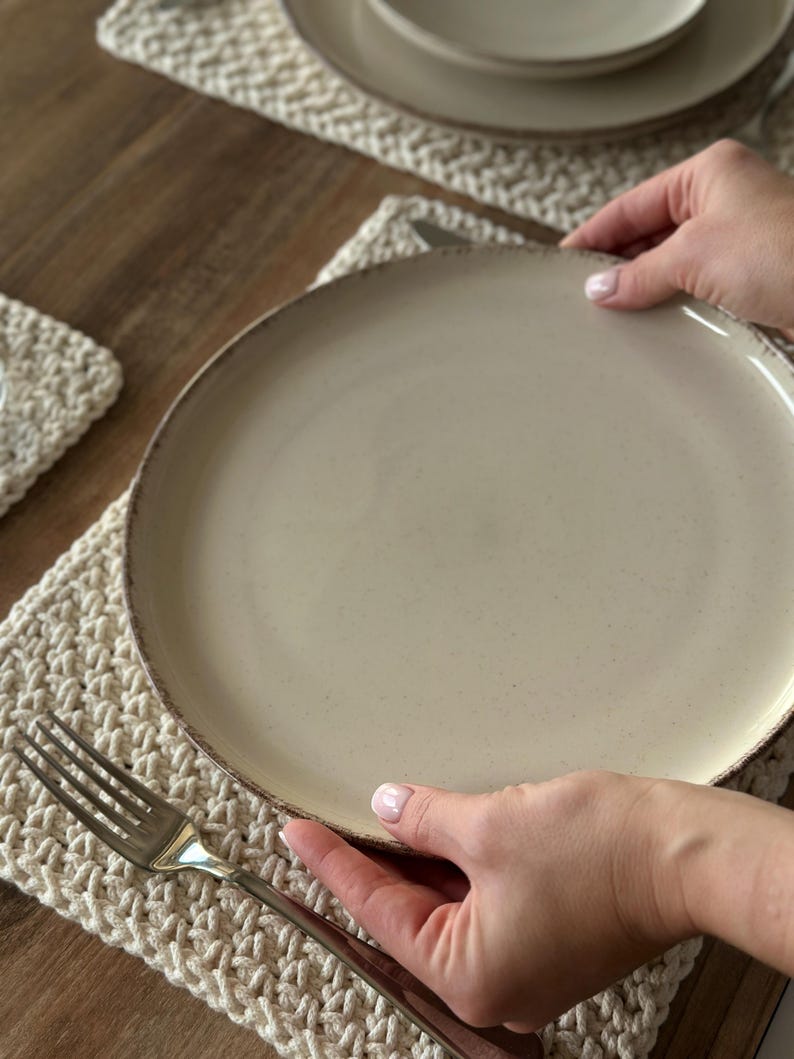 May include: A beige ceramic plate with a speckled surface and a dark brown rim is held by two hands. A silver fork rests on a cream-colored crocheted placemat. Additional plates and a bowl are visible in the background, suggesting a dining setting.