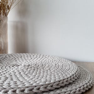 May include: Two round, light gray placemats crafted from thick, braided cord. The placemats feature a dense, circular woven pattern with a distinct braided edge, stacked one on top of the other to display the textured, handmade construction.