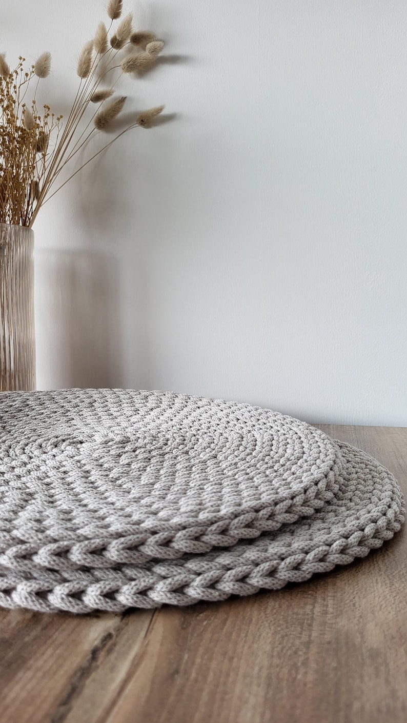May include: Two round, light gray placemats crafted from thick, braided cord. The placemats feature a dense, circular woven pattern with a distinct braided edge, stacked one on top of the other to display the textured, handmade construction.