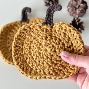 May include: Two crocheted pumpkin-shaped coasters in a mustard yellow color with brown stems. The coasters are made with a textured stitch.