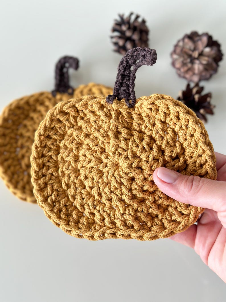 May include: Two crocheted pumpkin-shaped coasters in a mustard yellow color with brown stems. The coasters are made with a textured stitch.