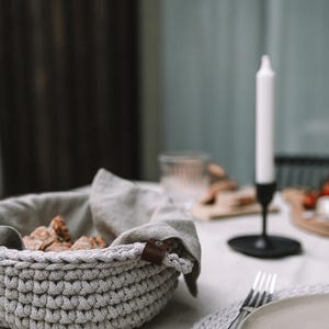 以下が含まれることがあります： 白いテーブルの上に、茶色の革タグが付いたグレーの編み込みバスケットが置かれています。バスケットの縁にはリネン製のナプキンがかけられています。バスケットの中にはパンが入っています。背景には白いキャンドルと黒い燭台が見えます。