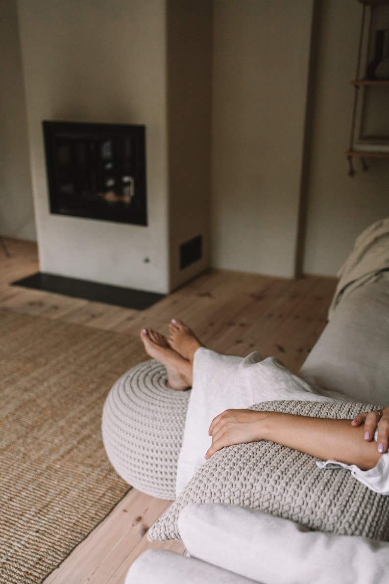 K&ouml;nnte beinhalten: Eine Person entspannt sich auf einem wei&szlig;en Sofa mit einem gestrickten Fu&szlig;hocker und einem Kissen. Das Sofa steht in einem Raum mit einem Kamin und Holzboden.