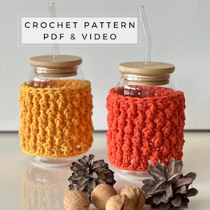 Op de afbeelding: Twee gehaakte glazen potten met een hoes, een oranje en een gele, met houten deksels en heldere glazen rietjes. De hoezen zijn gemaakt met een textuur en liggen op een wit oppervlak met dennenappels en walnoten.