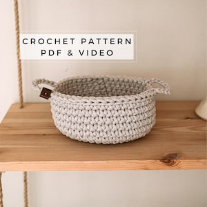 Könnte beinhalten: Ein gehäkelter Korb mit zwei Henkeln aus hellgrauem Garn. Der Korb steht auf einem Holzregal. Der Text "CROCHET PATTERN PDF & VIDEO" ist über dem Korb zu sehen.