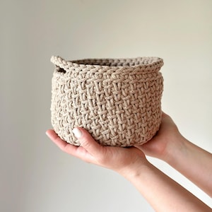 May include: A beige crocheted basket with a woven pattern. The basket is being held by two hands.