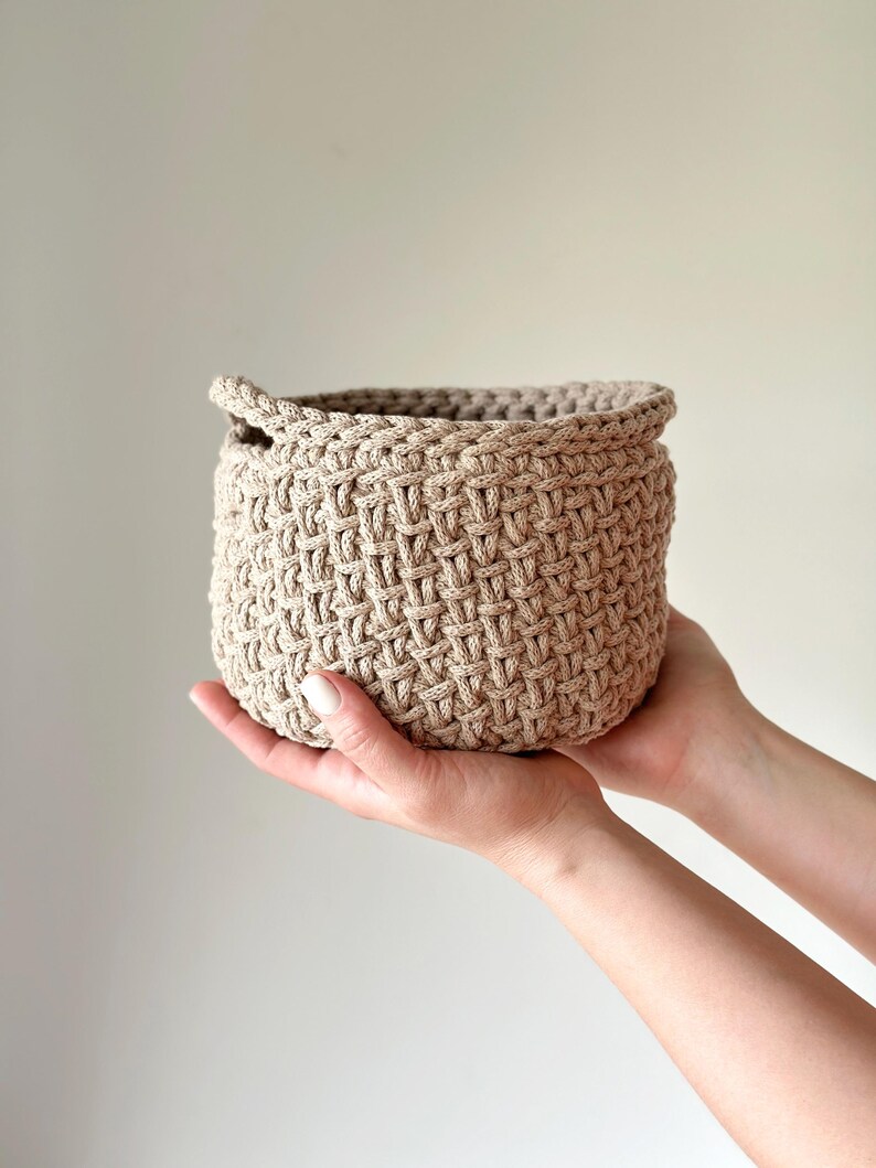 May include: A beige crocheted basket with a woven pattern. The basket is being held by two hands.