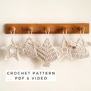May include: Five off-white crochet ornaments in star, angel, and tree shapes hang from a wooden rack. The ornaments have a textured appearance. The text "CROCHET PATTERN PDF & VIDEO" is displayed below.