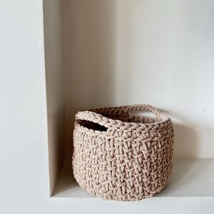 May include: A beige crocheted basket with two handles. The basket is sitting on a white shelf.