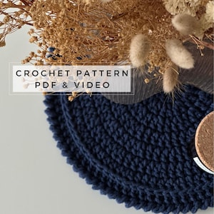 May include: A navy blue crochet coaster with a textured design. The coaster is on a white surface with dried flowers in the background. The text "CROCHET PATTERN PDF & VIDEO" is displayed on the image.