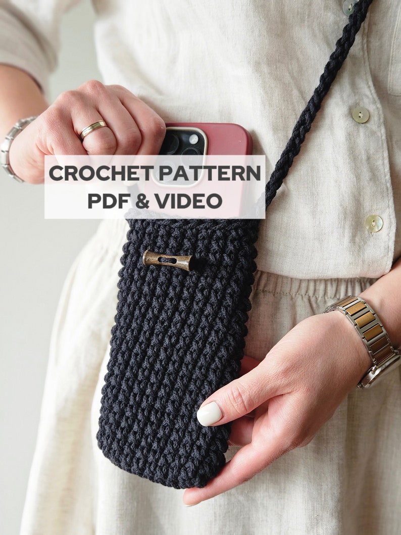 Op de afbeelding: Een zwarte gehaakte telefoonhoes met een bruine metalen sluiting. De hoes is bevestigd aan een zwarte touwband. De tekst "CROCHET PATTERN PDF & VIDEO" wordt weergegeven op een wit rechthoek.