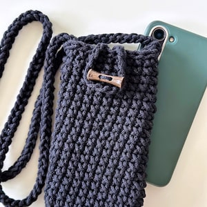 Op de afbeelding: Een zwarte gehaakte telefoonhoes met een houten knoopsluiting en een lange zwarte band. De hoes ligt op een wit oppervlak naast een groene telefoonhoes.