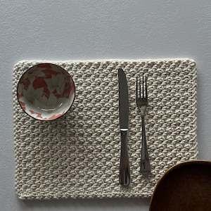 May include: A cream-colored, rectangular crocheted placemat with a bowl, knife, fork, and wooden bowl. The bowl has a floral pattern in shades of orange and white. The silverware is stainless steel.