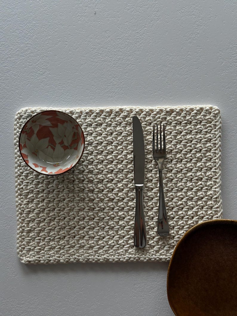May include: A cream-colored, rectangular crocheted placemat with a bowl, knife, fork, and wooden bowl. The bowl has a floral pattern in shades of orange and white. The silverware is stainless steel.