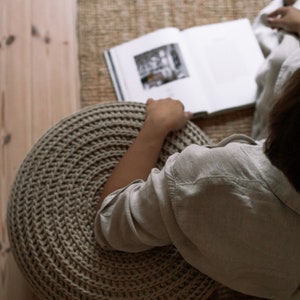 K&ouml;nnte beinhalten: Eine Person sitzt auf einem gro&szlig;en, runden, beigefarbenen Strickhocker und liest ein Buch. Der Hocker ist aus dickem, strukturiertem Garn gefertigt und hat ein kreisf&ouml;rmiges Muster.