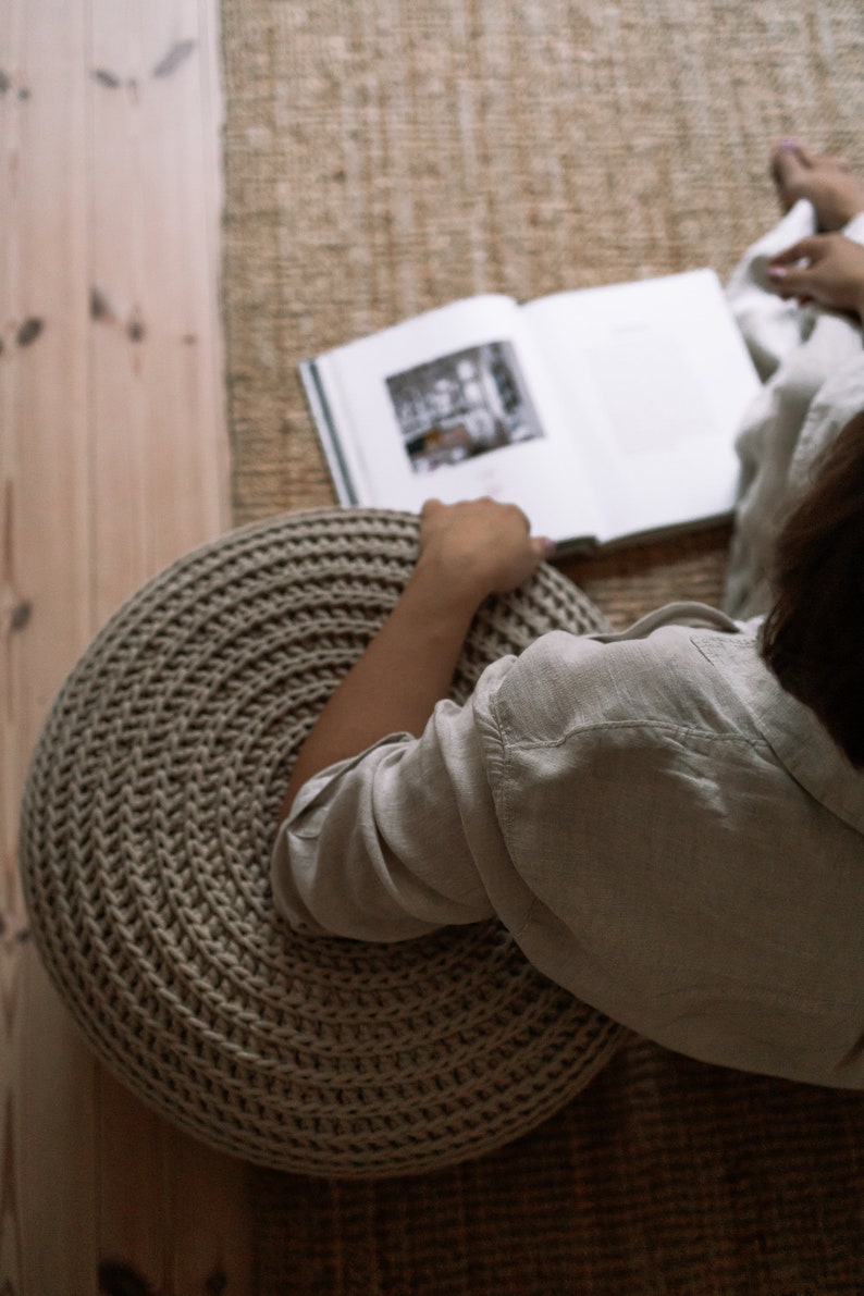 K&ouml;nnte beinhalten: Eine Person sitzt auf einem gro&szlig;en, runden, beigefarbenen Strickhocker und liest ein Buch. Der Hocker ist aus dickem, strukturiertem Garn gefertigt und hat ein kreisf&ouml;rmiges Muster.