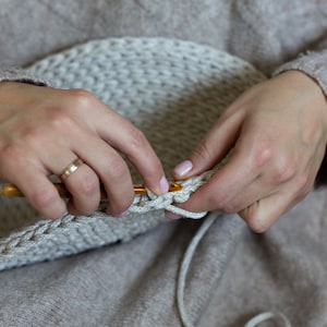 May include: Close-up of a person's hands crocheting with a beige yarn and a wooden crochet hook. The person is wearing a beige sweater.