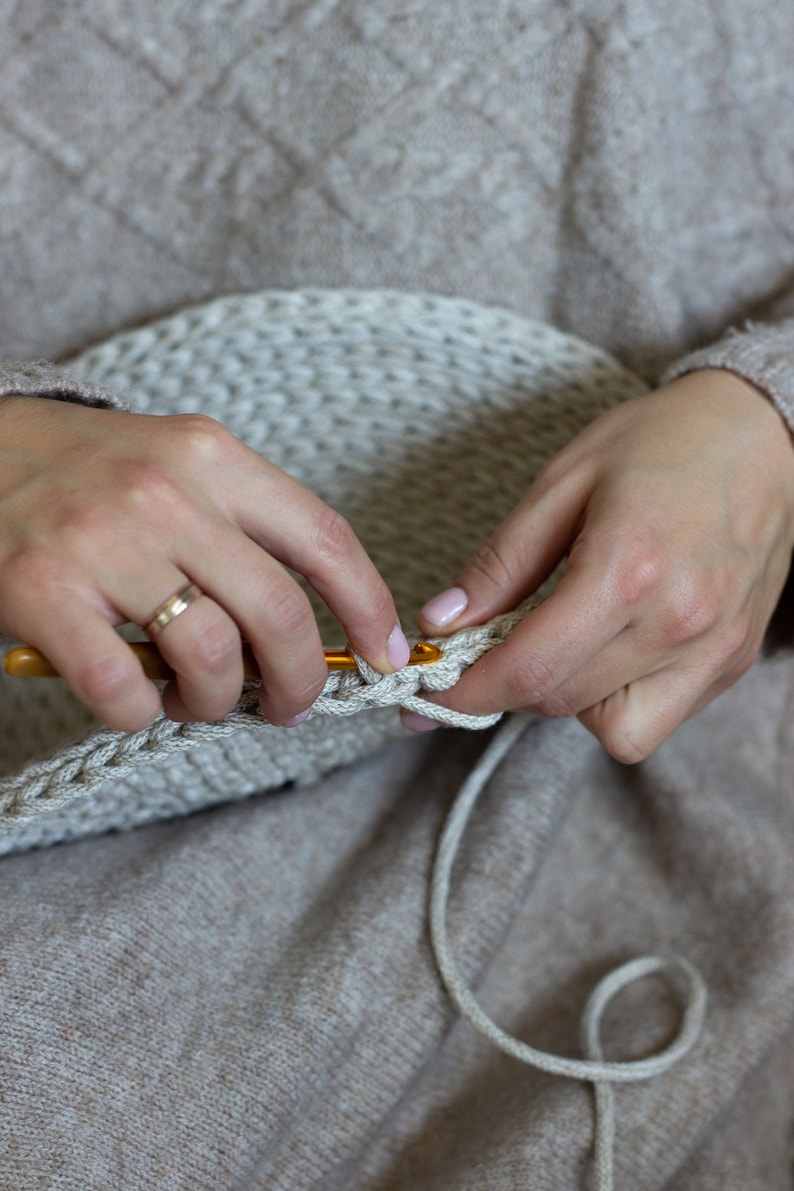 May include: Close-up of a person's hands crocheting with a beige yarn and a wooden crochet hook. The person is wearing a beige sweater.