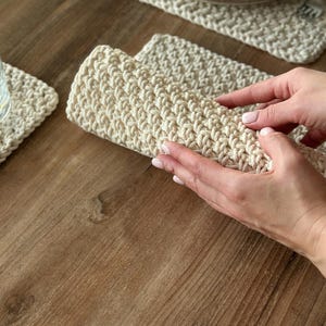 May include: Cream-colored, hand-crocheted placemats and coasters. The image shows a set of rectangular placemats and square coasters with a textured, woven appearance. The placemat is being held up, showing the thickness of the material. A plate and glass are visible.