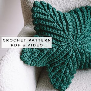Könnte beinhalten: Ein tealgrünes, gehäkeltes Kissen in Sternform auf einem weißen, flauschigen Stuhl. Der Text "CROCHET PATTERN PDF & VIDEO" ist auf dem Bild zu sehen.