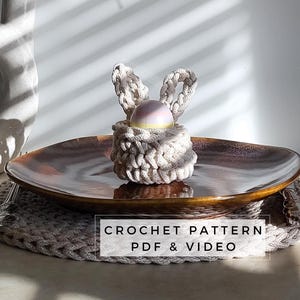 Pode incluir: Um ninho de ovo de crochê em forma de coelho com um ovo rosa pastel e amarelo dentro. O ninho está sobre um prato marrom com um tapete de crochê embaixo. O texto "CROCHET PATTERN PDF & VIDEO" é exibido abaixo do ninho.