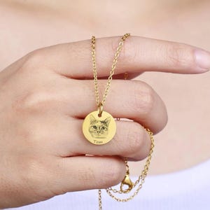 May include: A gold-coloured necklace with a circular pendant. The pendant has an engraved cat portrait and the name "Tom". The delicate chain is held by a person's hand.