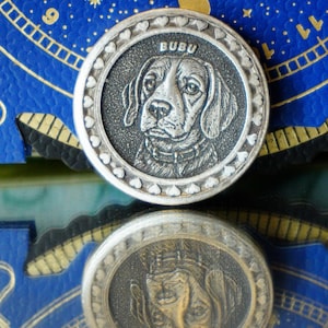 May include: A silver-colored round pendant with a detailed engraving of a dog's face, the name "BUBU" above the dog. The pendant has a border of small hearts. The pendant is reflected in a shiny surface.