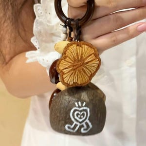 May include: A mushroom-themed keychain featuring a brown wooden ring, a woven mushroom cap, and a stone-like base with a white graphic. The keychain has a brown cord and small beads. The mushroom cap is a light brown color.