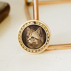 May include: A gold-colored pendant featuring a detailed engraving of a cat's face. The circular pendant has a border of small hearts and is suspended from a thin, gold-toned chain. The cat's portrait is in a sepia tone.