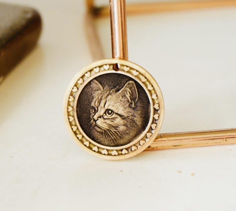 May include: A gold-colored pendant featuring a detailed engraving of a cat's face. The circular pendant has a border of small hearts and is suspended from a thin, gold-toned chain. The cat's portrait is in a sepia tone.